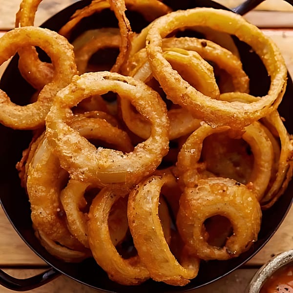 A close-up of golden-brown, crispy onion rings in a dark bowl, with a dipping sauce visible.